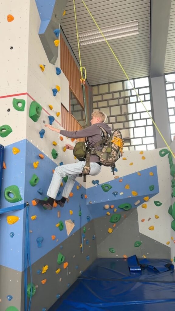 Ein Mann mit Rucksack klettert an einer Indoor-Kletterwand und greift nach einem blauen Griff; Sicherheitsmatten und bunte Griffe sind zu sehen.