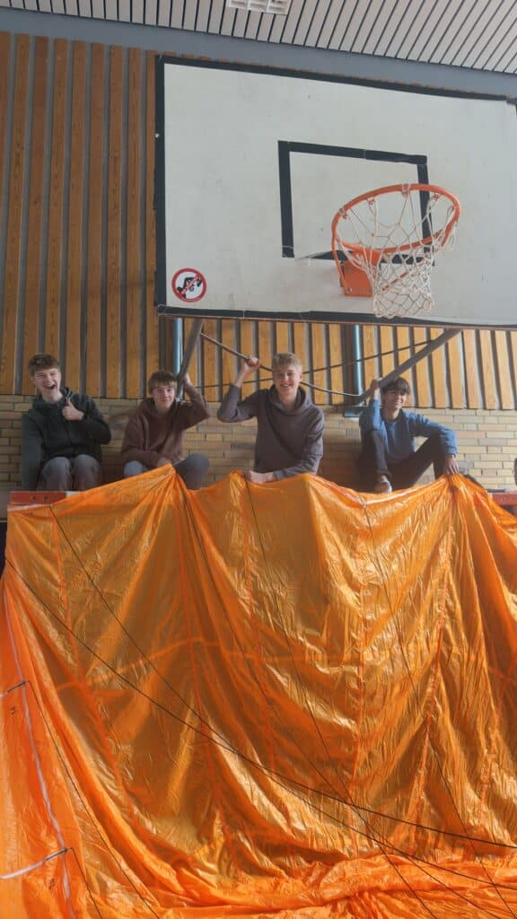 Vier junge Leute sitzen auf einer Tribüne in einer Turnhalle und halten Seile an einem großen orangefarbenen Fallschirm, der vor ihnen unter einem Basketballkorb gespannt ist.