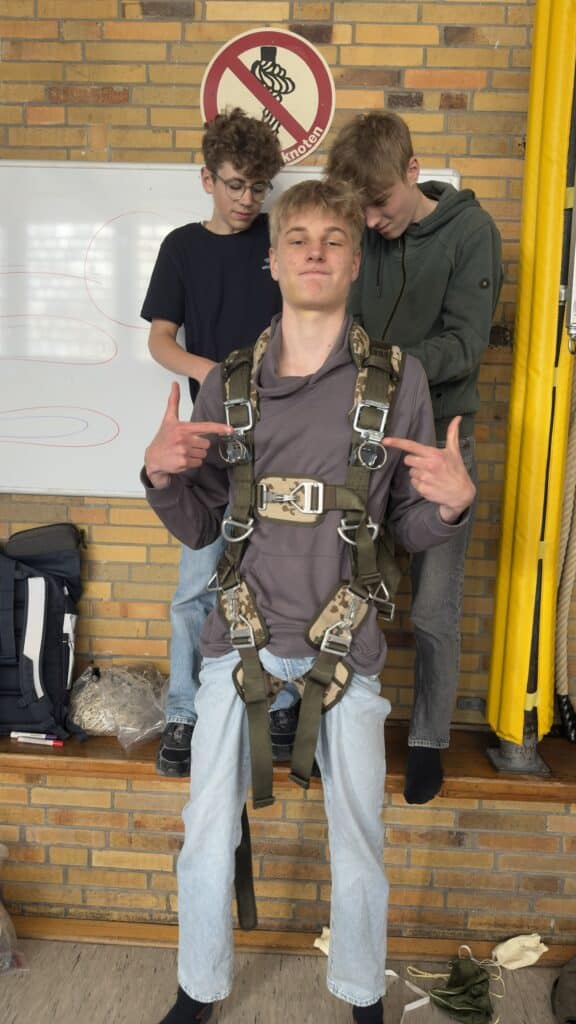 Ein junger Mann mit einem Fallschirmgurt sitzt auf einer Bank und deutet auf sich selbst, während zwei andere hinter ihm stehen und die Gurte einstellen.