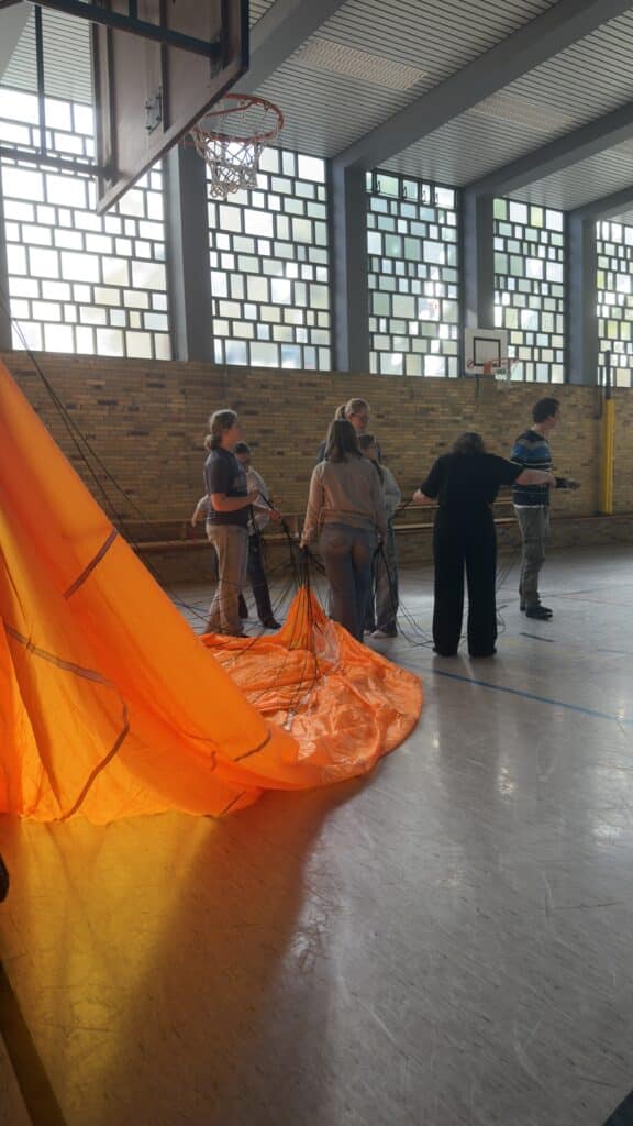 Eine Gruppe von Menschen steht in einer Turnhalle und hält Seile an einem großen, leuchtend orangefarbenen Fallschirm, der auf dem Boden ausgebreitet ist.