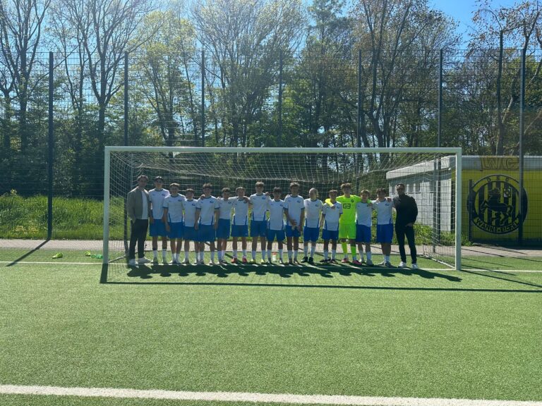 Eine Fußballmannschaft in weißen und blauen Trikots steht gemeinsam vor einem Tor auf einem sonnigen Feld im Freien mit Bäumen und einem gelben Gebäude im Hintergrund.