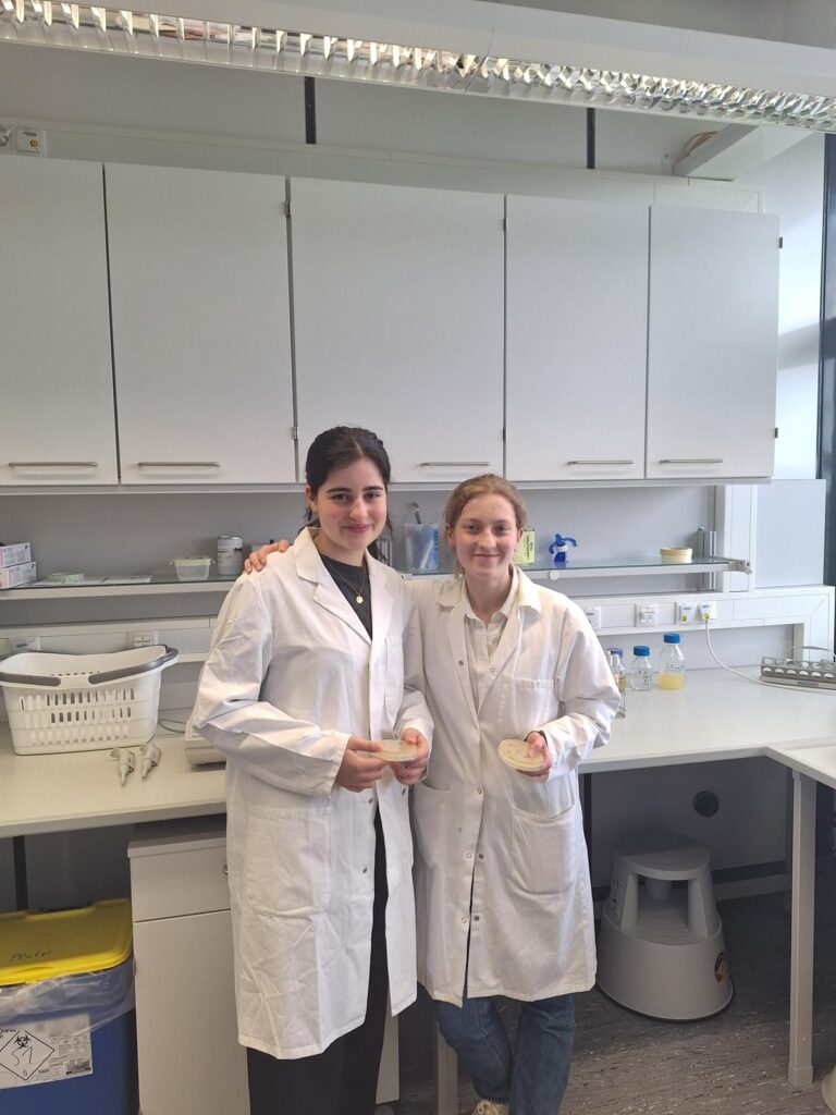 Zwei Personen in weißen Laborkitteln stehen in einem Labor, lächeln und halten Petrischalen. Im Hintergrund sind Laborgeräte und Schränke zu sehen.