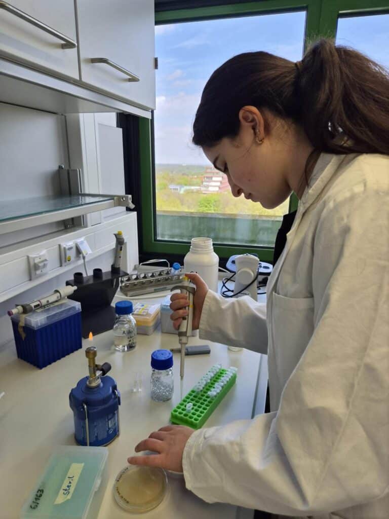 Eine Frau im Laborkittel pipettiert in einem Labor Flüssigkeit in Reagenzgläser. Im Hintergrund sind Laborgeräte und ein Fenster mit Blick ins Grüne zu sehen.
