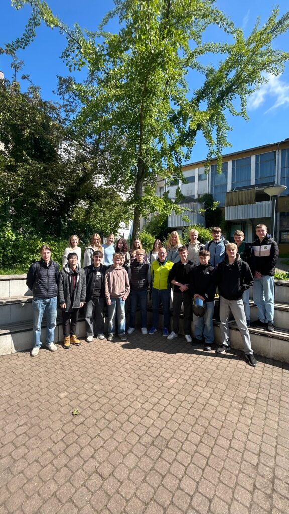Eine Gruppe von 19 Personen steht an einem sonnigen Tag im Freien vor einem belaubten Baum und modernen Gebäuden und posiert und lächelt für ein Gruppenfoto.