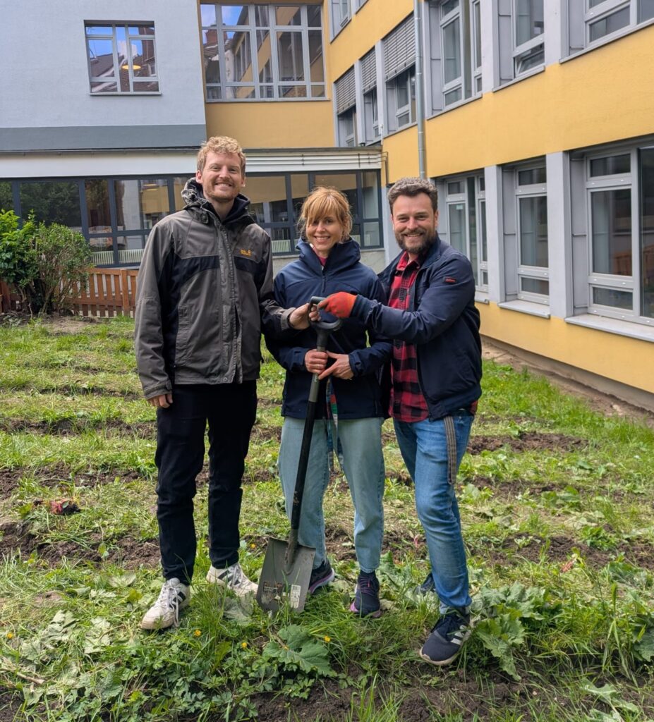 Drei Menschen stehen lächelnd in einem grasbewachsenen Hof, einer hält eine Schaufel, im Hintergrund ein gelb-graues Gebäude.