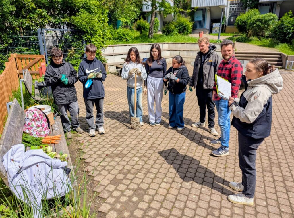 Eine Gruppe von acht Personen steht in einem Halbkreis auf einer gepflasterten Fläche im Freien, trägt Handschuhe und legere Kleidung und bereitet sich auf eine Garten- oder Aufräumaktion vor.
