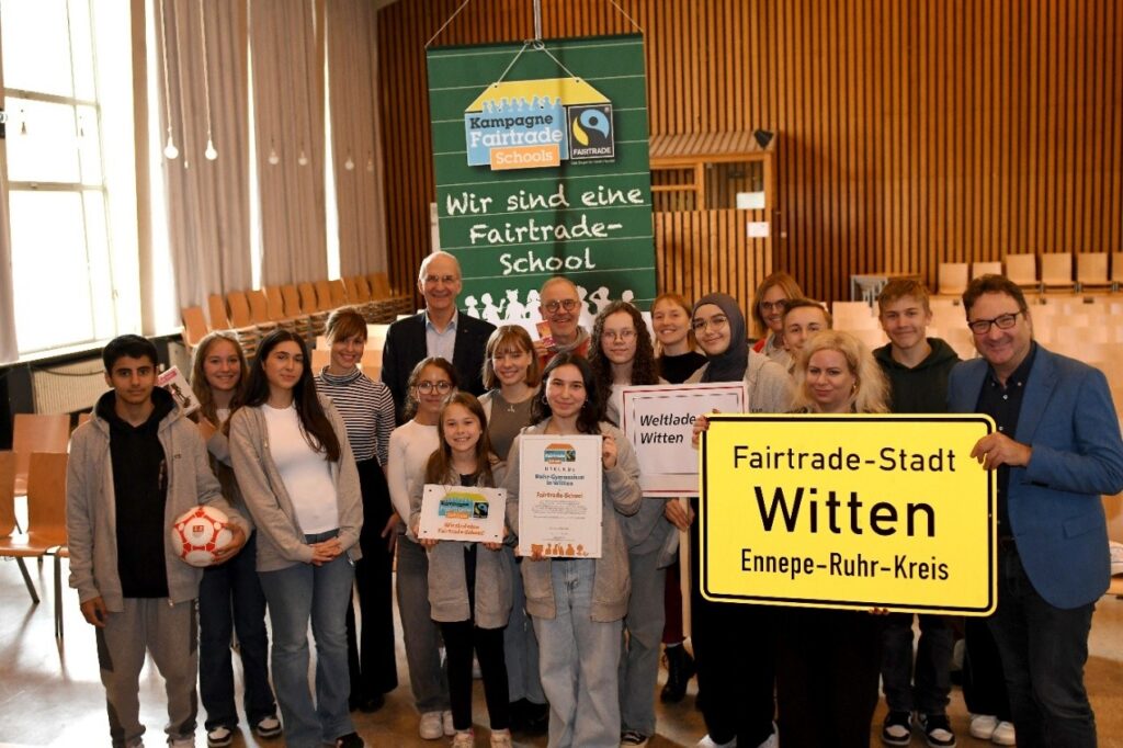 Eine Gruppe von Schülern und Erwachsenen posiert mit Schildern mit der Aufschrift Fairtrade-Stadt Witten und Wir sind eine Fairtrade-School in einer Halle mit Stühlen.