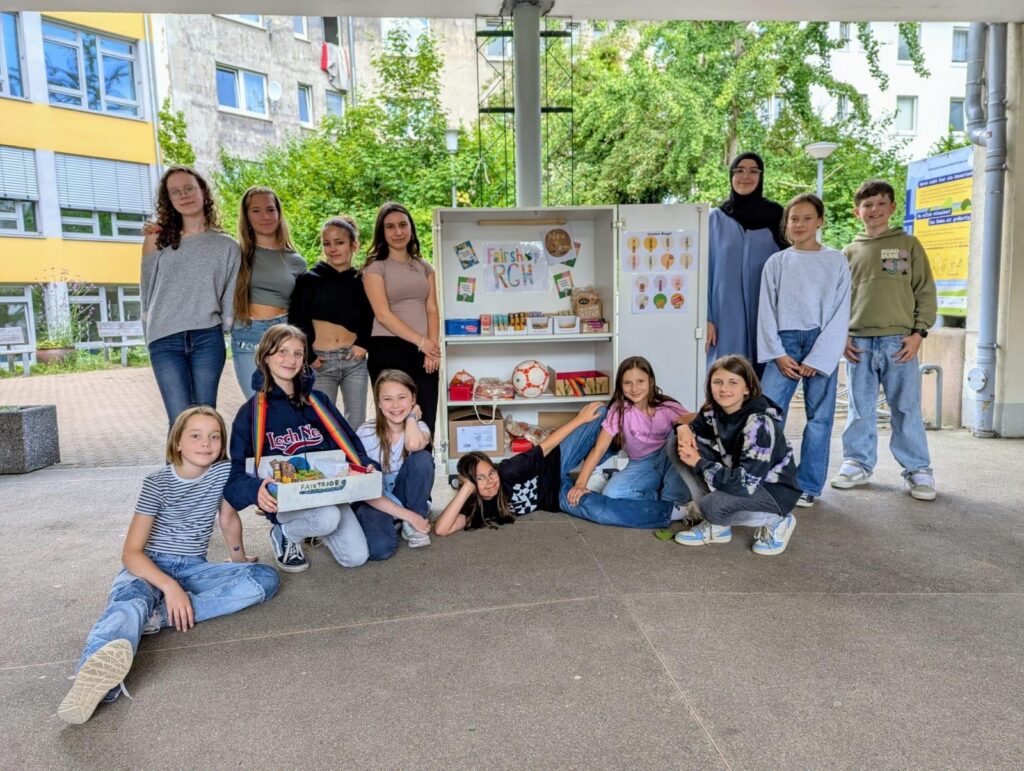 Eine Gruppe von Jugendlichen und Kindern posiert vor einem Gebäude um ein Regal mit Büchern, Bastelmaterial und einem Fußball und lächelt in die Kamera.