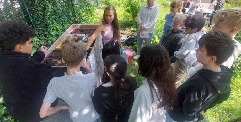 Eine Gruppe von Kindern versammelt sich um eine Frau, die in einer üppigen, grünen Umgebung Gartenarbeit oder Kompostierung mit Behältern demonstriert.