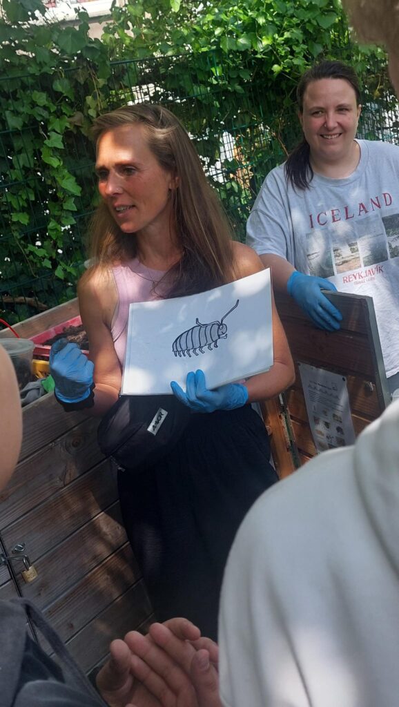 Eine Frau mit blauen Handschuhen hält eine Zeichnung eines Pillenkäfers hoch, während sie zu einer Gruppe von Menschen spricht. Eine andere Frau lächelt im Hintergrund.