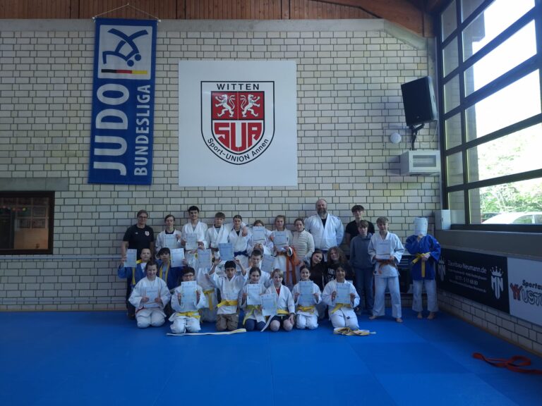 Eine Gruppe von Judoka posiert in einer Halle auf blauen Matten, mit Urkunden in der Hand, mit Trainern im Hintergrund und einem Banner der Judo-Bundesliga an der Wand.