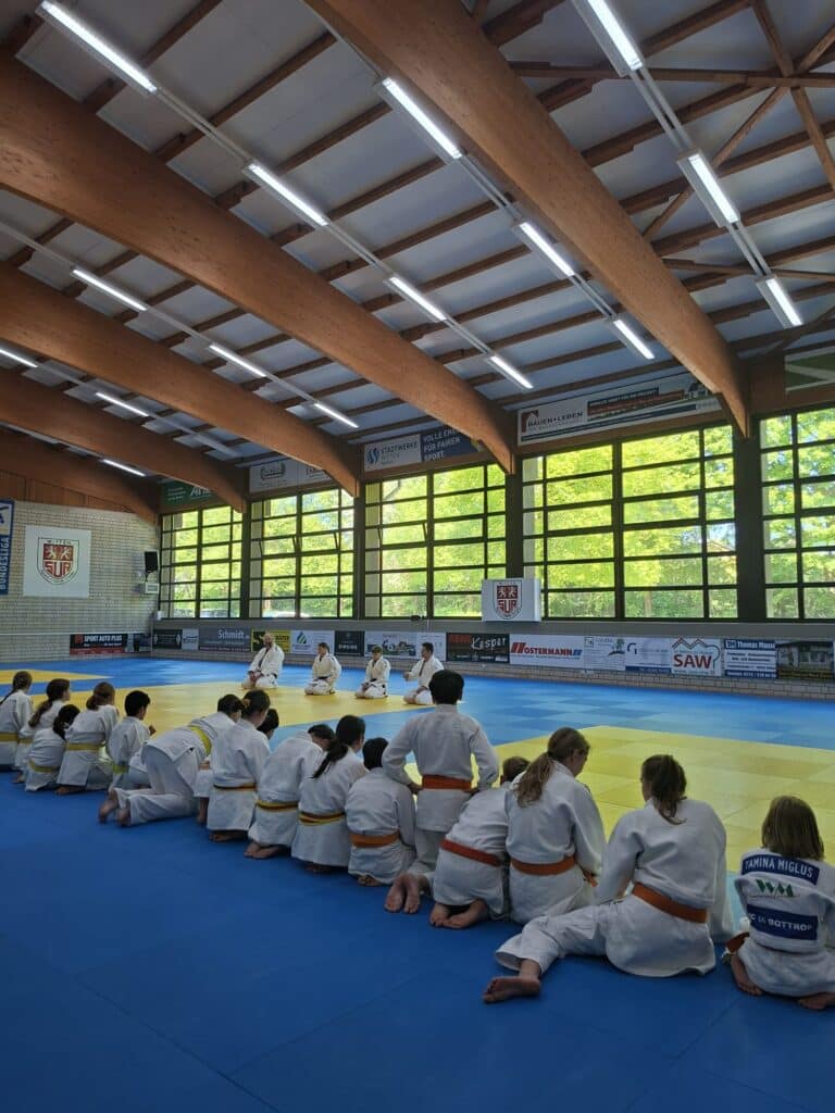 Eine Gruppe von Kindern in Judo-Uniformen sitzt auf einer blauen Matte und sieht zu, wie ein Lehrer in einer hell erleuchteten Turnhalle mit großen Fenstern Techniken vorführt.