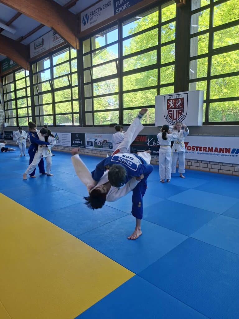 Judosportler üben Wurftechniken in einer hell erleuchteten Turnhalle mit großen Fenstern und gelb-blauen Matten. Andere Sportler schauen zu oder trainieren im Hintergrund.