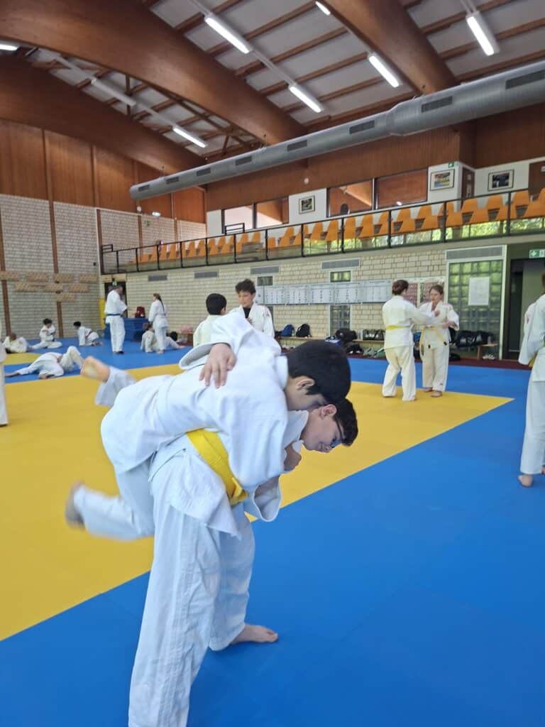 Kinder in weißen Judoanzügen üben Würfe auf einer blau-gelben Matte in einer großen Turnhalle mit Holzdecke und Zuschauern im Hintergrund.