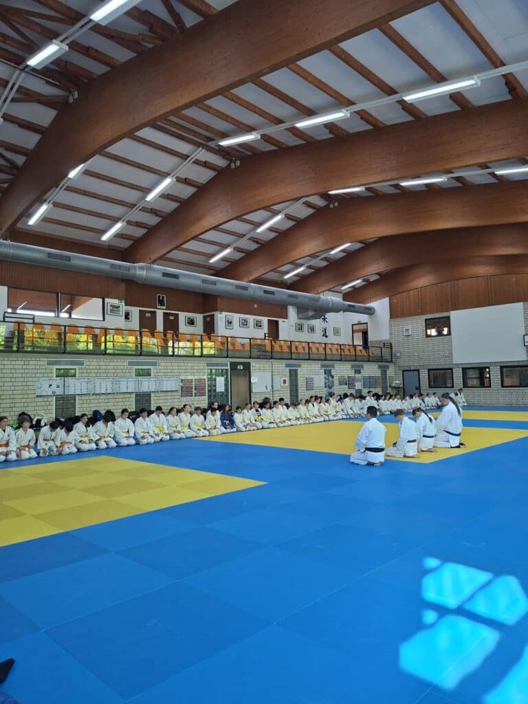 Eine Gruppe von Menschen in weißen Uniformen sitzt in Reihen auf blauen und gelben Matten in einem großen Dojo mit Holzbalken und hohen Decken.