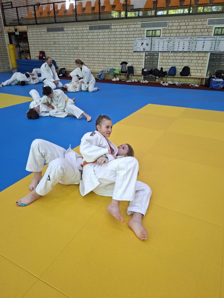 Zwei Mädchen in weißen Judoanzügen üben eine Grifftechnik auf einer gelben Matte, während andere im Hintergrund eines Dojos trainieren.