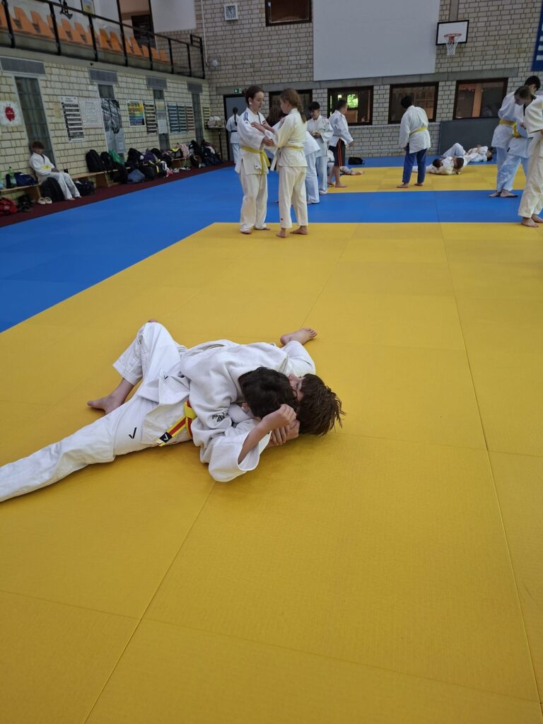Zwei Kinder in weißen Judoanzügen üben einen Haltegriff auf einer gelben Matte, während andere in ähnlicher Kleidung im Hintergrund trainieren.