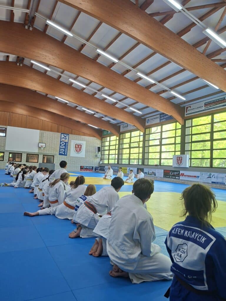 Kinder in weißen Judoanzügen knien in Reihen auf Matten in einer hellen Turnhalle, mit Blick auf die Ausbilder an der Vorderseite unter großen Fenstern und Holzbalken.