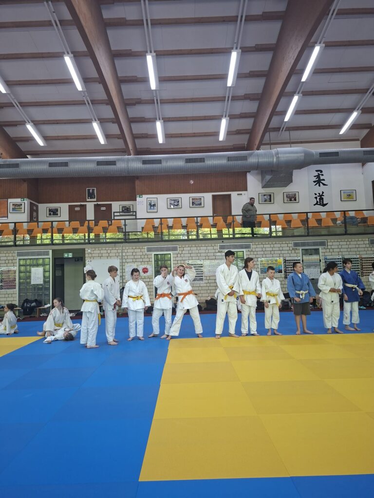 Eine Gruppe von Personen in weißen Judoanzügen mit gelben Gürteln steht in einer Reihe auf einer blau-gelben Matte in einer Turnhalle mit leeren Sitzreihen.
