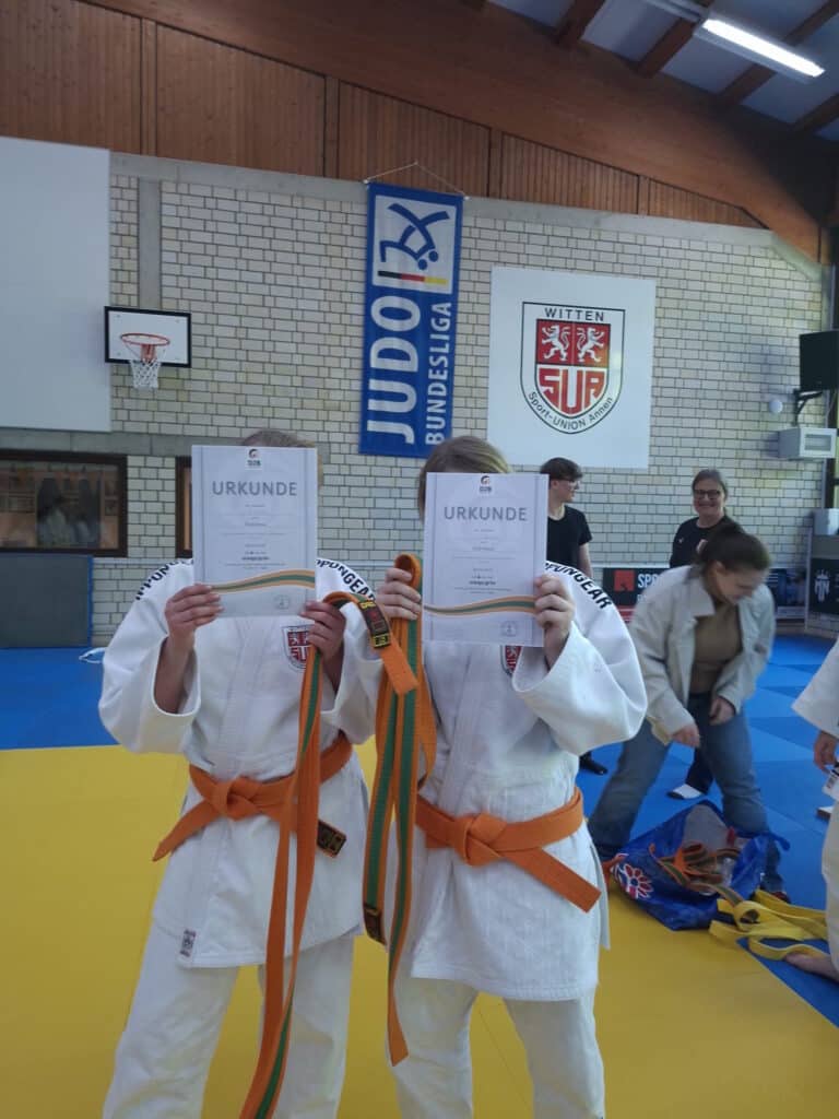Zwei Judosportler in weißen Uniformen und orange-grünen Gürteln halten sich in einer Turnhalle Urkunden vor das Gesicht, während im Hintergrund andere Sportler und Sportbanner zu sehen sind.