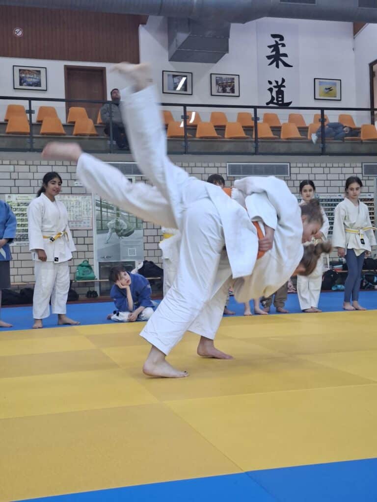Zwei Mädchen in weißen Judoanzügen kämpfen auf einer Matte, wobei die eine die andere wirft. Andere Kinder in Judoanzügen schauen im Hintergrund zu.