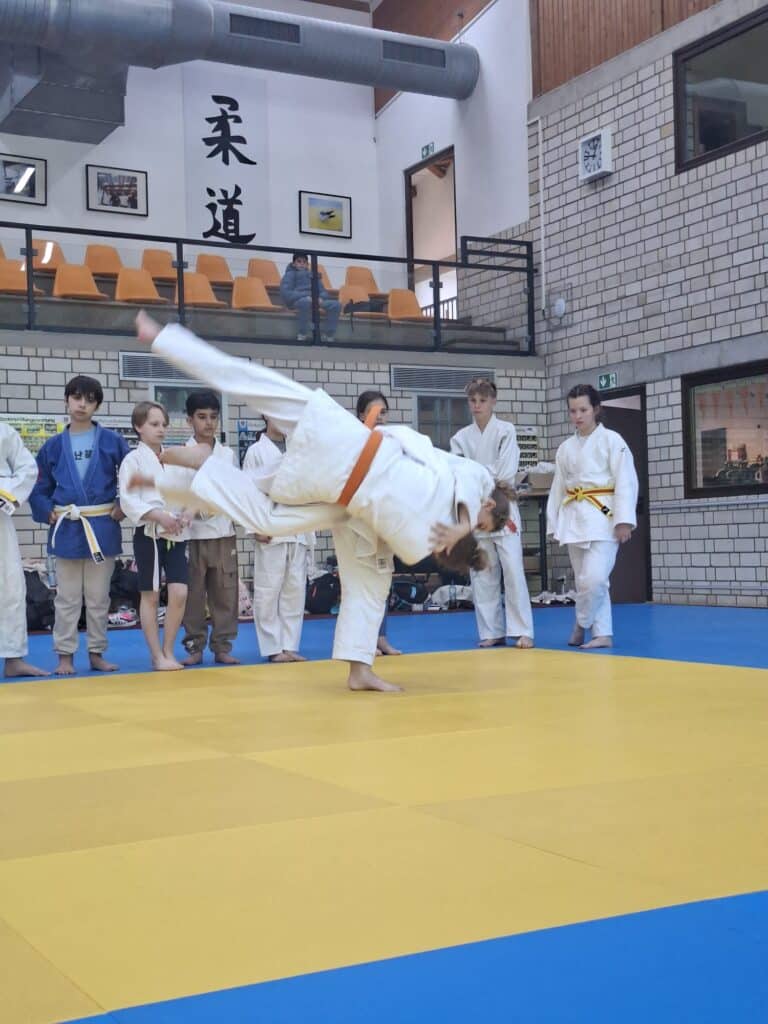 Zwei junge Judosportler in weißen Uniformen kämpfen, wobei der eine den anderen über die Hüfte stößt, während mehrere Kinder auf einer Matte in einem Dojo zusehen.