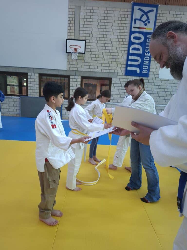 Kinder in Judoanzügen erhalten von Erwachsenen Urkunden und gelbe Gürtel in einer Turnhalle mit einem JUDO-Banner und einem Basketballkorb im Hintergrund.