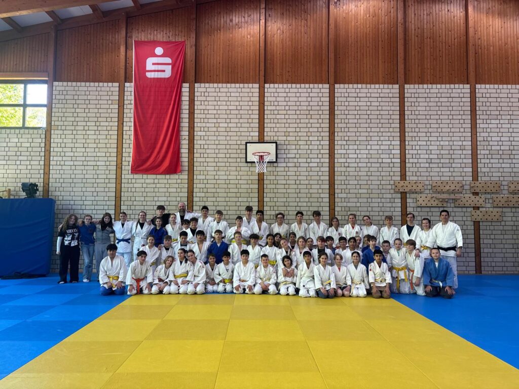 Eine große Gruppe von Kindern und Erwachsenen in Judo-Uniformen posiert gemeinsam in einer Turnhalle mit blauen und gelben Matten, Backsteinwänden und einem roten Banner, das über ihnen hängt.