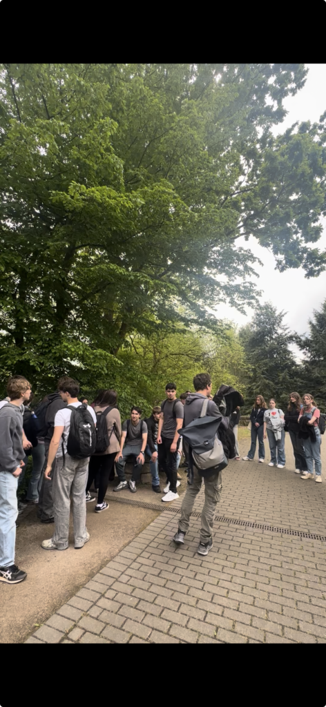 Eine Gruppe von Menschen mit Rucksäcken steht und unterhält sich auf einem gepflasterten Weg, umgeben von grünen Bäumen und Grünpflanzen in einem Park.