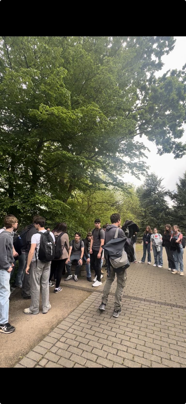 Eine Gruppe von Menschen mit Rucksäcken steht und unterhält sich auf einem gepflasterten Weg, umgeben von grünen Bäumen und Grünpflanzen in einem Park.