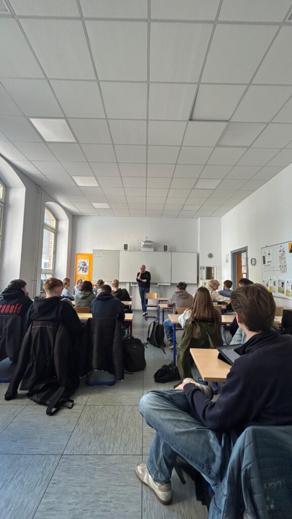 Ein Lehrer steht vorne in einem Klassenzimmer und spricht zu den Schülern, die an den Tischen sitzen, mit Fenstern auf der linken Seite und einer Tafel hinter dem Lehrer.