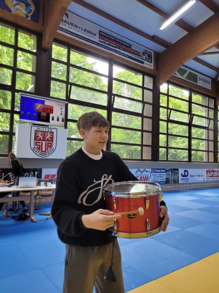 Ein junger Mann in einem schwarzen Pullover hält eine rote Trommel in einer Sporthalle mit blauen und gelben Matten und großen Fenstern.
