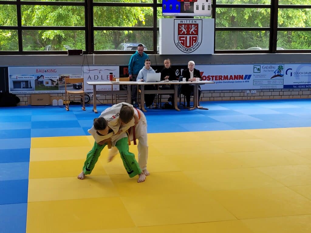 Zwei junge Judoka kämpfen auf einer gelb-blauen Matte, während Schiedsrichter und Kampfrichter an einem Tisch im Hintergrund in einer Sporthalle sitzen.