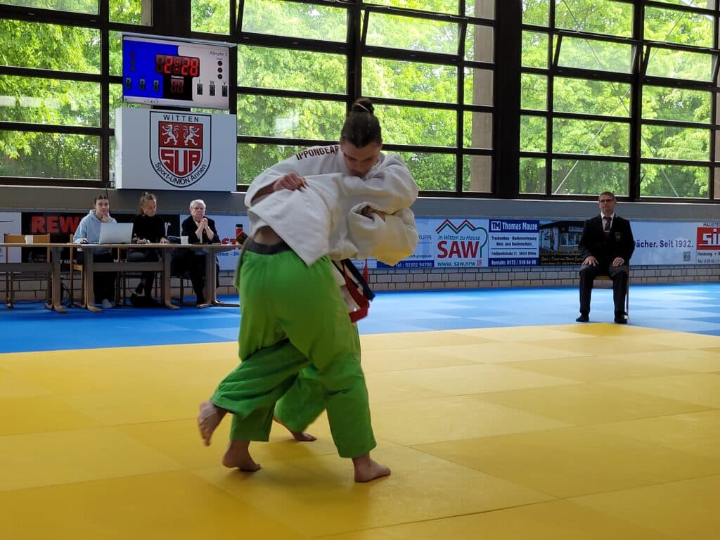 Zwei Judosportler kämpfen während eines Wettkampfs auf einer gelben Matte, während die Kampfrichter an einem Tisch sitzen und große Fenster im Hintergrund zu sehen sind.