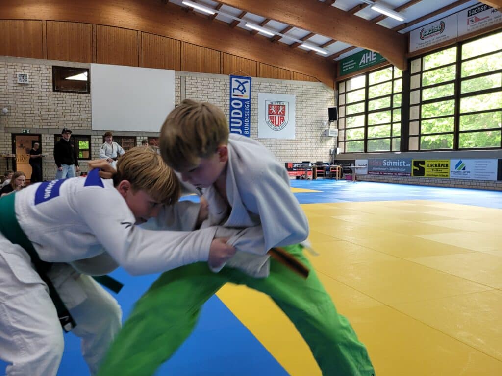 Zwei Jungen in Judoanzügen kämpfen auf einer gelb-blauen Matte in einer Sporthalle und konzentrieren sich während eines Judokampfes.