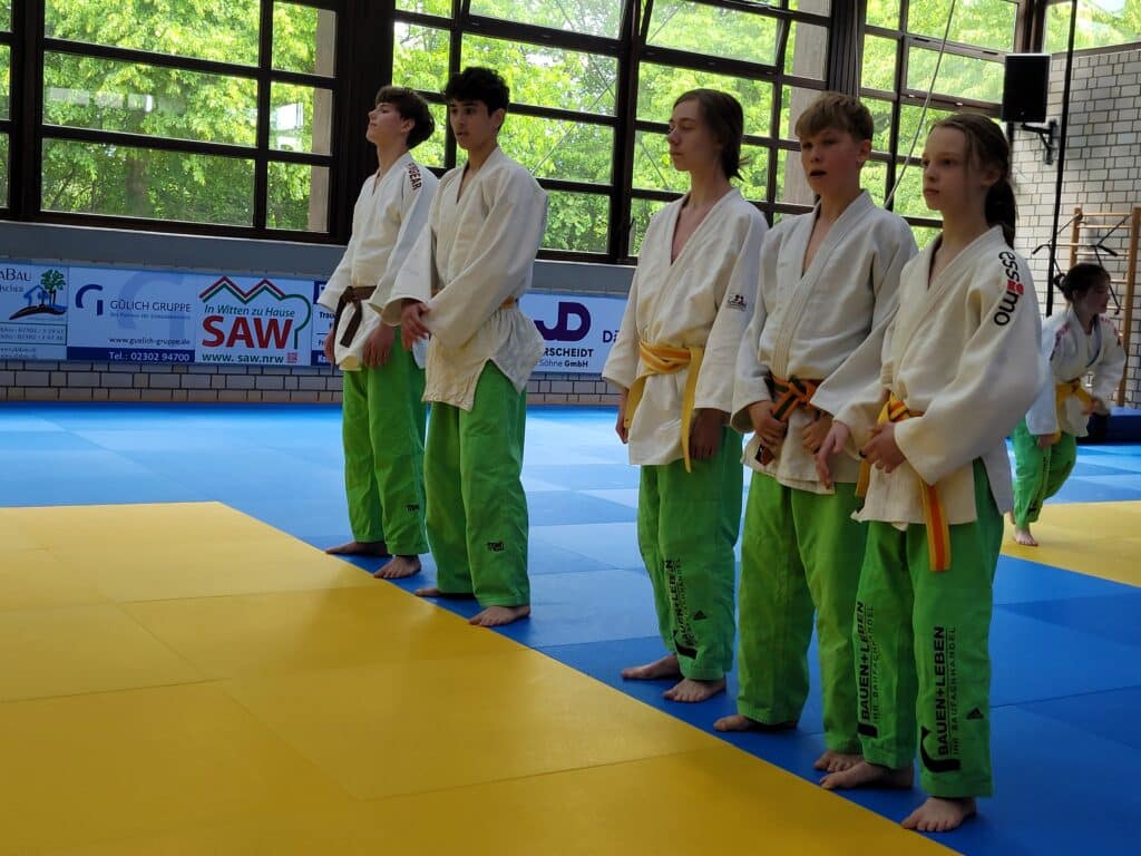 Fünf junge Leute in weißen Judoanzügen und grünen Hosen stehen barfuß in einer Reihe auf einer blau-gelben Matte in einer Turnhalle mit großen Fenstern.