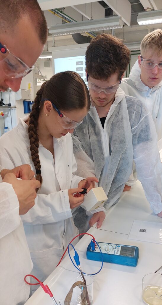 Vier Studenten in Laborkitteln und Schutzbrillen arbeiten gemeinsam in einem Labor und prüfen mit Kabeln und einem Multimeter ein kleines Gerät, das auf einem Tisch steht.