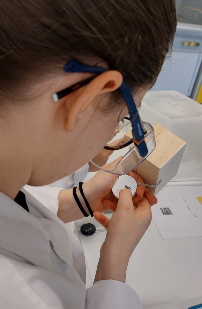 Eine Person, die eine Schutzbrille trägt, untersucht in einem Labor mit einer Lupe ein kleines weißes Objekt aus nächster Nähe.