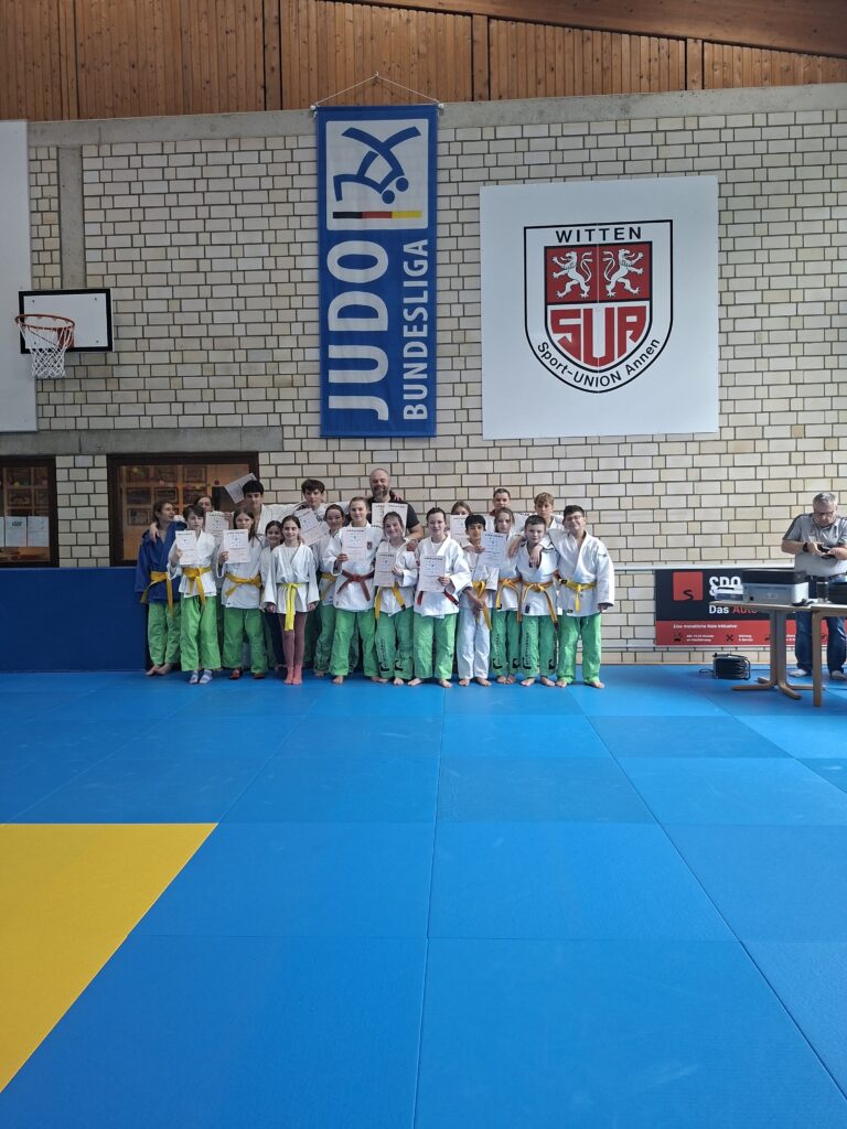 Eine Gruppe junger Judoschüler in weißen Jacken und grünen Hosen steht in einer Turnhalle zusammen, hält Gürtel, im Hintergrund sind Banner und eine Anzeigetafel zu sehen.