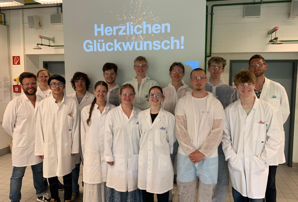 Eine Gruppe von Menschen in Laborkitteln und Schutzbrillen steht lächelnd in einem Labor. Hinter ihnen ist auf einem Bildschirm die Aufschrift „Herzlichen Glückwunsch!“ auf Deutsch zu sehen.