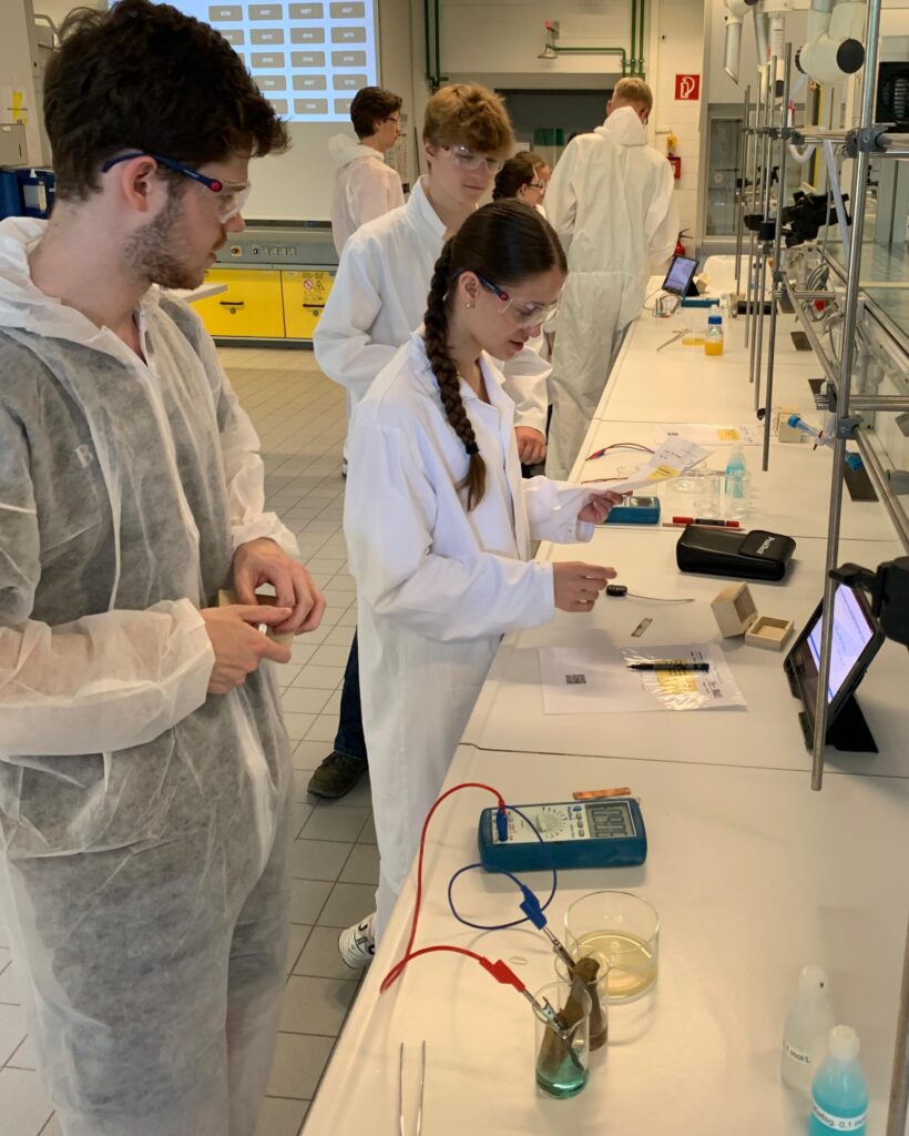Vier Personen in Laborkitteln und Schutzbrillen arbeiten an einem Labortisch mit wissenschaftlichen Geräten, Unterlagen und einem Tablet in einem hell beleuchteten Labor.