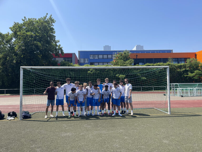 Eine Gruppe von Fußballspielern in blauen Shorts und weißen Hemden posiert an einem sonnigen Tag gemeinsam vor einem Tor auf einem Feld im Freien.