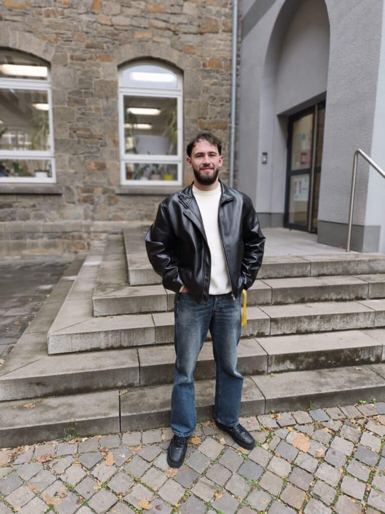 Ein bärtiger Mann in schwarzer Lederjacke und Jeans steht auf einer Steintreppe vor einem Gebäude mit großen Fenstern und einem gewölbten Eingang.
