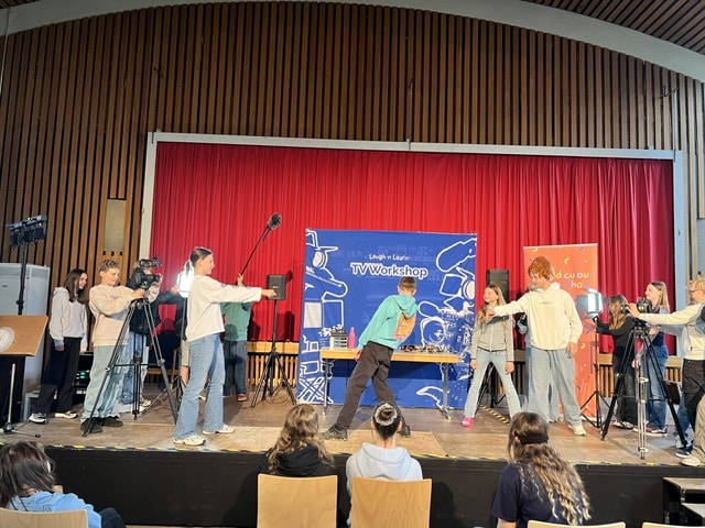 Eine Gruppe junger Leute filmt während eines Fernseh-Workshops eine Szene auf der Bühne – mit Kameras, Mikrofonen und Requisiten –, während das Publikum zusieht.