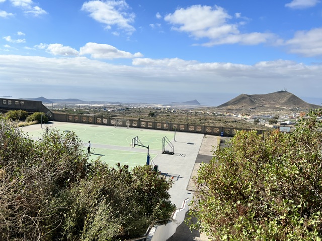 Ein von Grünflächen umgebener Basketballplatz im Freien, im Hintergrund die Berge und ein strahlend blauer Himmel an einem sonnigen Tag.