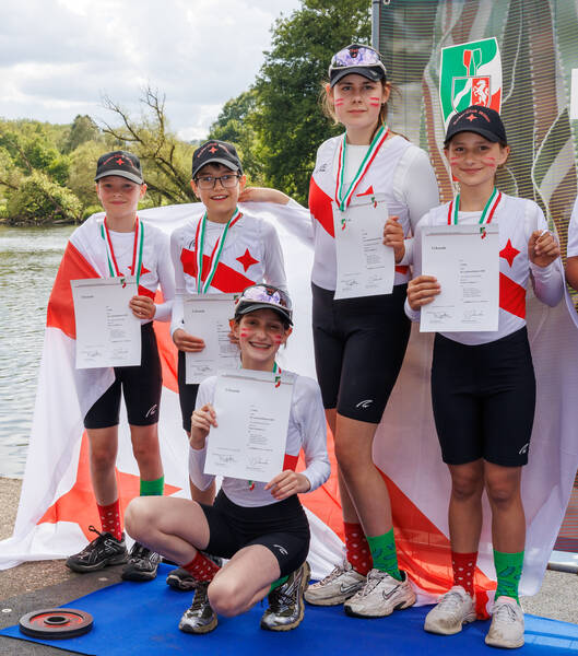 Fünf junge Ruderer mit Medaillen und in einheitlichen Trikots halten Urkunden in den Händen und lächeln für ein Foto am Wasser, um ihren Erfolg zu feiern.