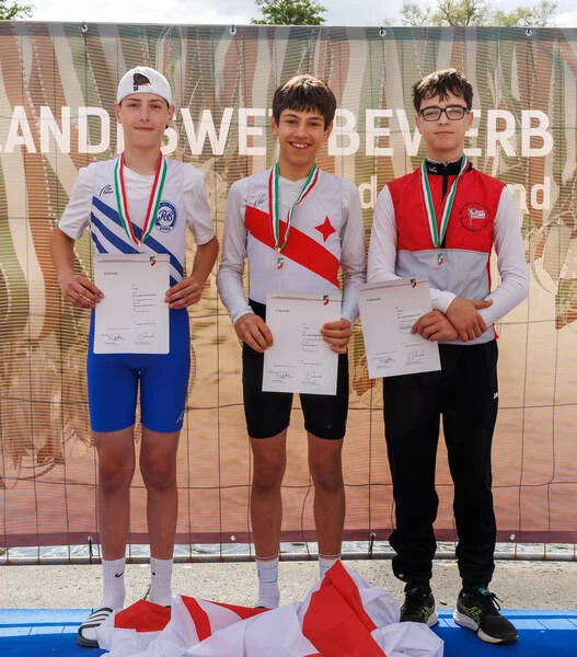 Drei Teenager in Radfahrerkleidung stehen auf einem Podium, halten Urkunden in den Händen, tragen Medaillen um den Hals und lächeln in die Kamera.