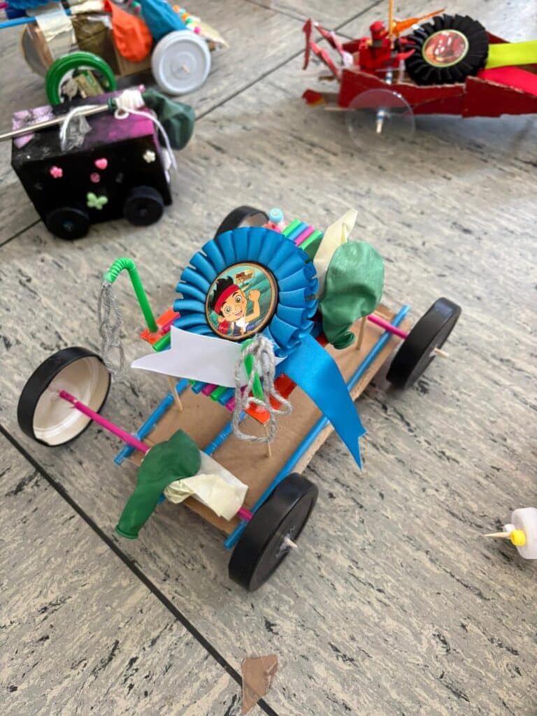 Ein handgefertigtes Spielzeugauto mit Rädern, Luftballons, Strohhalmen und einer blauen Schleife, auf der ein Comic-Kind abgebildet ist, steht auf einem Fliesenboden inmitten anderer bunter Spielzeugautos.