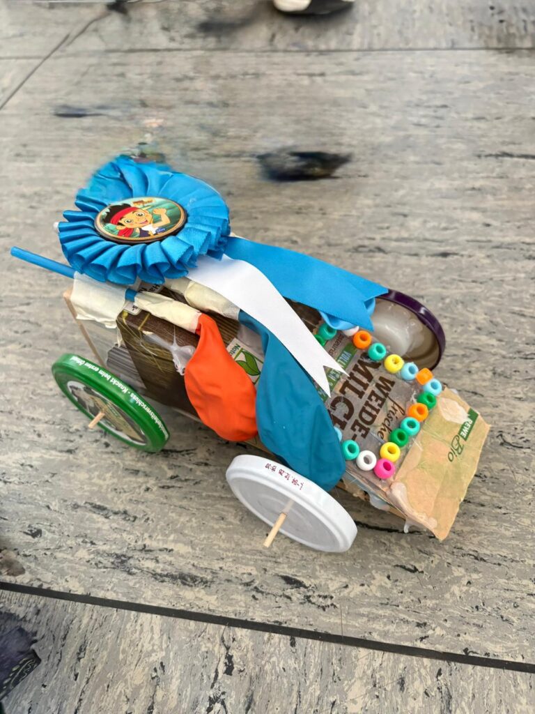 Ein selbstgebautes Spielzeugauto aus recycelten Materialien, mit Flaschenverschlüssen als Rädern, aus Pappe, Plastik und Perlen, verziert mit Luftballons und einer blauen Schleife als Auszeichnung oben drauf.