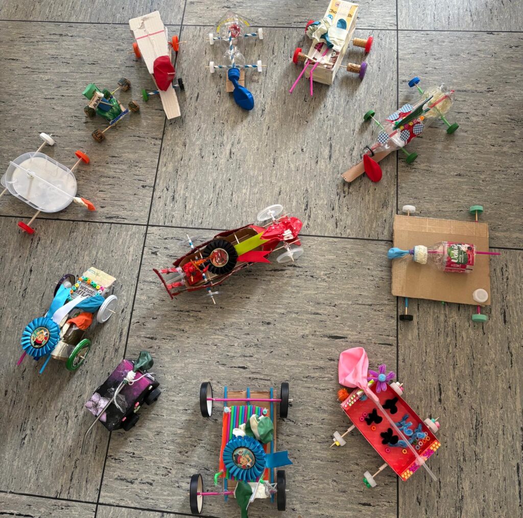 Ein Kreis aus selbstgebastelten Spielzeugautos aus recycelten Materialien, Luftballons und Flaschenverschlüssen, die auf einem grauen Fliesenboden angeordnet sind.